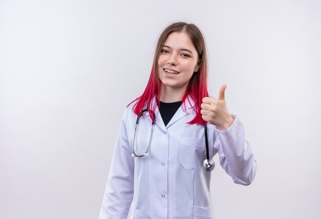 Sonriente joven médico con estetoscopio bata médica su pulgar hacia arriba sobre fondo blanco aislado