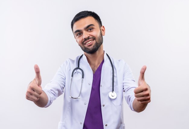 Sonriente joven médico con estetoscopio bata médica sus pulgares hacia arriba sobre fondo blanco aislado