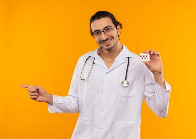 Sonriente joven médico con gafas médicas vistiendo bata médica con estetoscopio sosteniendo pastillas y apunta a un lado en amarillo