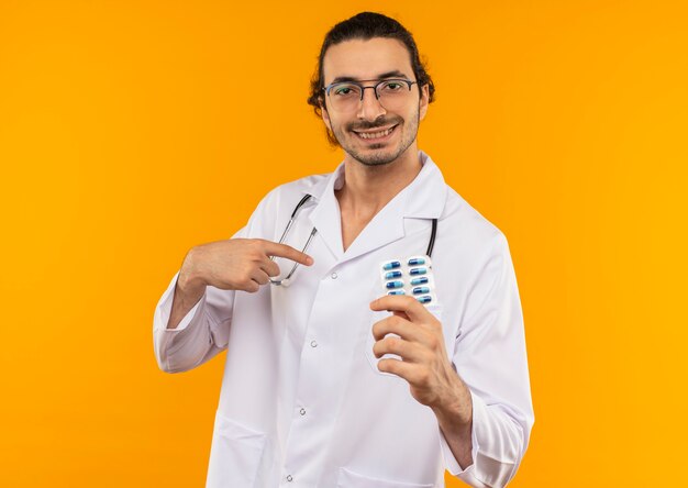 Sonriente joven médico con gafas médicas vistiendo bata médica con estetoscopio sosteniendo pastillas y apunta a sí mismo