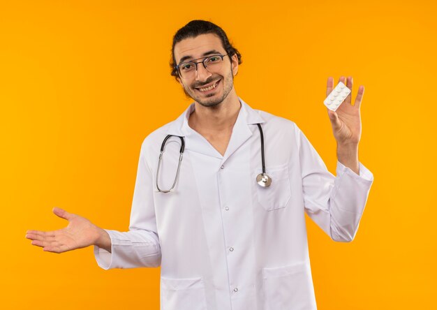 Sonriente joven médico con gafas médicas vistiendo bata médica con estetoscopio sosteniendo píldoras y puntos con la mano al lado en amarillo