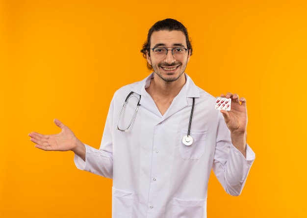 Sonriente joven médico con gafas médicas vistiendo bata médica con estetoscopio sosteniendo píldoras puntos con la mano al lado en la pared amarilla aislada con espacio de copia
