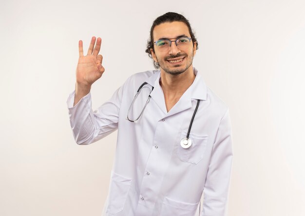 Sonriente joven médico con gafas ópticas vistiendo bata blanca con estetoscopio mostrando gesto okey en pared blanca aislada con espacio de copia