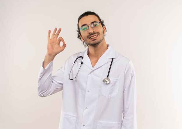 Sonriente joven médico con gafas ópticas vistiendo bata blanca con estetoscopio mostrando gesto okey en pared blanca aislada con espacio de copia