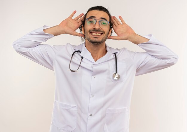 Sonriente joven médico con gafas ópticas vistiendo bata blanca con estetoscopio mostrando gesto de orejas de conejo en pared blanca aislada con espacio de copia