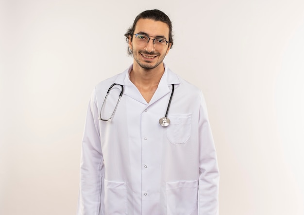Sonriente joven médico con gafas ópticas vistiendo bata blanca con estetoscopio en pared blanca aislada con espacio de copia