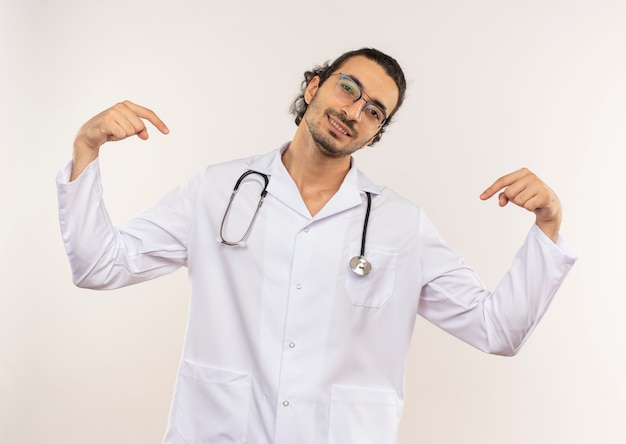 Sonriente joven médico con gafas ópticas vistiendo túnica blanca con estetoscopio apunta a sí mismo en blanco