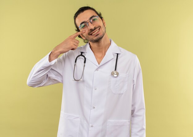 Sonriente joven médico con gafas ópticas vistiendo túnica blanca con estetoscopio apunta su ojo en verde