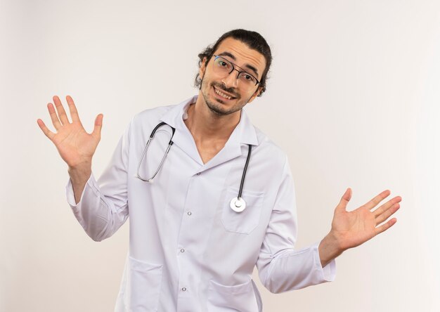 Sonriente joven médico con gafas ópticas vistiendo túnica blanca con estetoscopio extiende las manos sobre la pared blanca aislada con espacio de copia