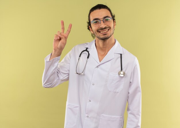 Sonriente joven médico con gafas ópticas vistiendo túnica blanca con estetoscopio mostrando gesto de paz en verde