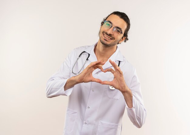 Sonriente joven médico con gafas ópticas vistiendo túnica blanca con estetoscopio que muestra el gesto del corazón en la pared blanca aislada con espacio de copia