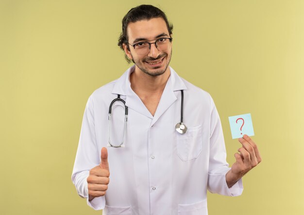 Sonriente joven médico con gafas ópticas vistiendo túnica blanca con estetoscopio sosteniendo papel signo de interrogación con el pulgar hacia arriba en la pared verde aislada con espacio de copia