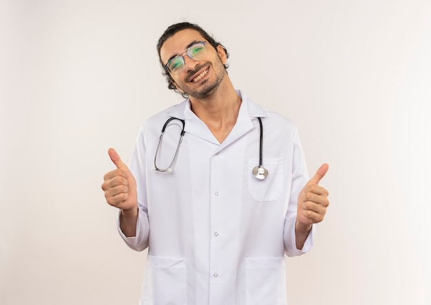 Foto gratuita sonriente joven médico con gafas ópticas vistiendo túnica blanca con estetoscopio sus pulgares hacia arriba en la pared blanca aislada con espacio de copia