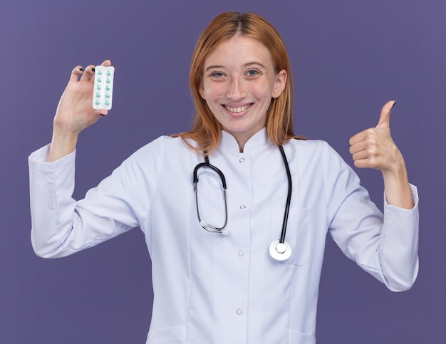 Sonriente joven médico de jengibre con bata médica y un estetoscopio que muestra el paquete de píldoras médicas a la cámara mostrando el pulgar hacia arriba aislado en la pared púrpura