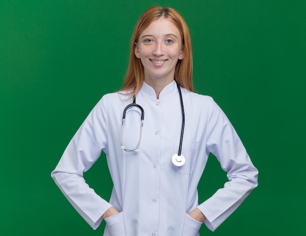 Sonriente joven médico de jengibre vistiendo bata médica y estetoscopio manteniendo las manos en los bolsillos