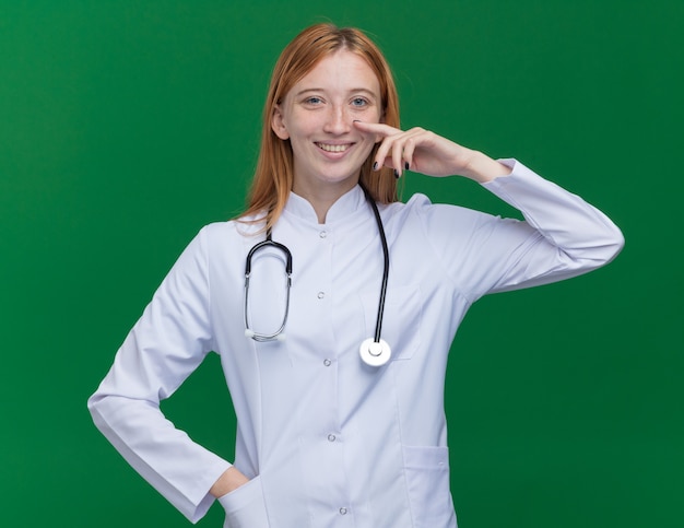 Sonriente joven médico de jengibre vistiendo bata médica y estetoscopio poniendo la mano en el bolsillo apuntando a su nariz aislada en la pared verde