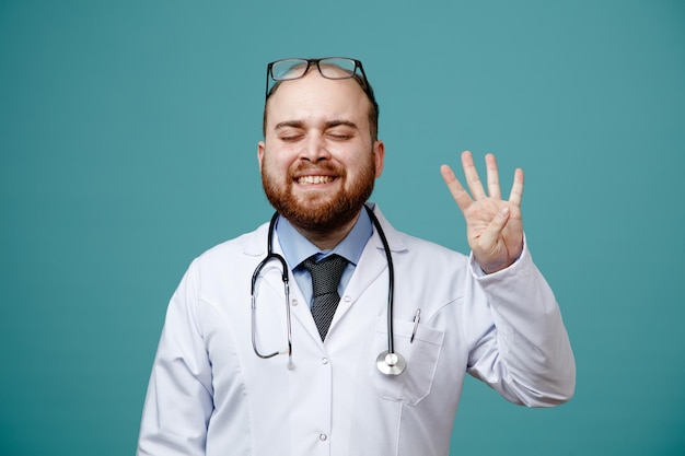 Sonriente joven médico masculino con abrigo médico y estetoscopio alrededor del cuello y anteojos en la cabeza que muestran cuatro con la mano con los ojos cerrados aislados en el fondo azul