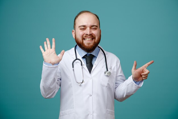 Sonriente joven médico masculino con abrigo médico y estetoscopio alrededor del cuello mirando a la cámara que muestra cinco con la mano apuntando al lado aislado en el fondo azul