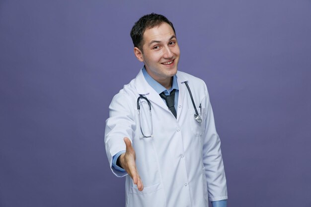 Sonriente joven médico masculino con bata médica y estetoscopio alrededor del cuello mirando a la cámara haciendo un gesto agradable para conocerte aislado en un fondo morado