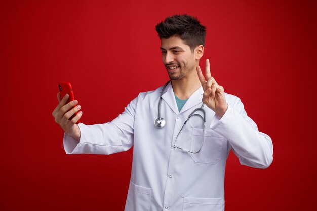 Sonriente joven médico masculino con uniforme médico y estetoscopio alrededor de su cuello estirando el teléfono móvil tomando selfie mostrando el signo de la paz aislado en el fondo rojo