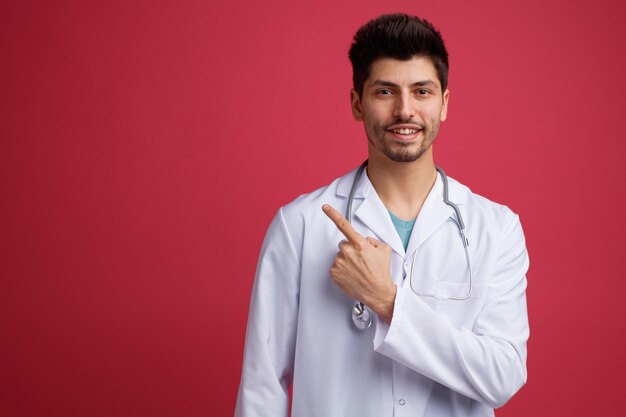 Sonriente joven médico masculino con uniforme médico y estetoscopio alrededor de su cuello mirando a la cámara apuntando al lado aislado en fondo rojo con espacio de copia