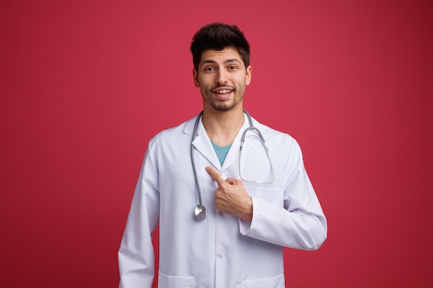 Sonriente joven médico masculino con uniforme médico y estetoscopio mirando a la cámara apuntándose a sí mismo aislado en el fondo rojo