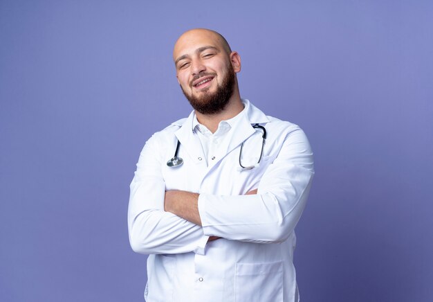 Sonriente joven médico varón calvo vistiendo bata médica y estetoscopio cruzando las manos aisladas sobre fondo azul.