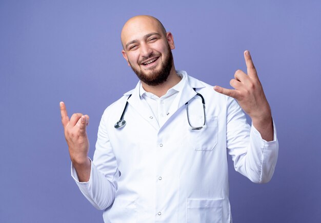 Sonriente joven médico varón calvo vistiendo bata médica y un estetoscopio mostrando gesto de cabra