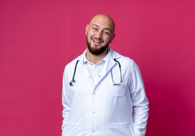 Sonriente joven médico varón calvo vistiendo bata médica y estetoscopio poniendo las manos en el bolsillo aislado en rosa con espacio de copia