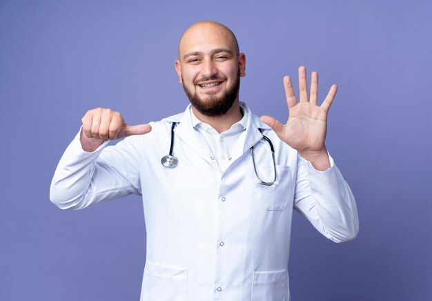 Sonriente joven médico varón calvo vistiendo bata médica y estetoscopio