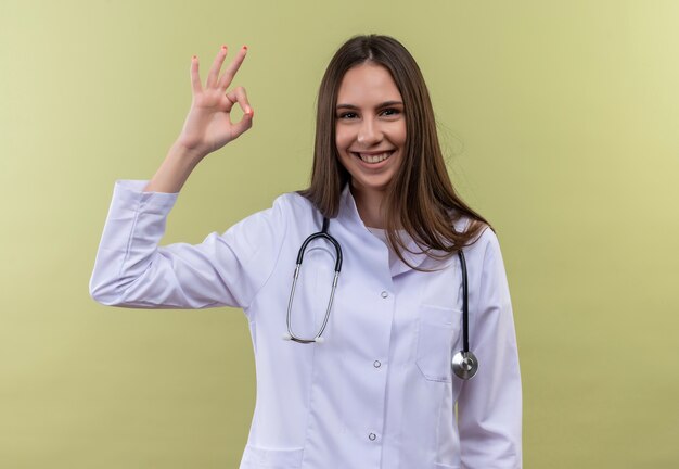 Sonriente joven médico vestida con estetoscopio bata médica mostrando gesto okey sobre fondo verde