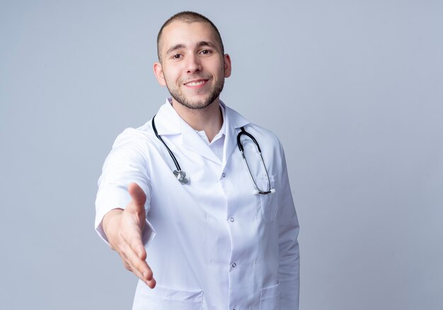 Sonriente joven médico vistiendo bata médica y un estetoscopio alrededor de su cuello estirando la mano al frente gesticulando hola aislado en la pared blanca