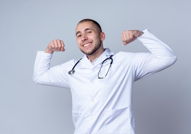 Sonriente joven médico vistiendo bata médica y un estetoscopio alrededor de su cuello gesticulando fuerte aislado en la pared blanca