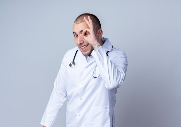 Sonriente joven médico vistiendo bata médica y un estetoscopio alrededor de su cuello haciendo gesto de mirada al frente aislado en la pared blanca