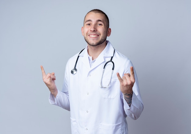 Sonriente joven médico vistiendo bata médica y un estetoscopio alrededor de su cuello haciendo signos de roca aislados en la pared blanca