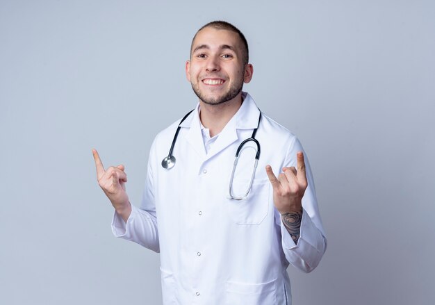 Sonriente joven médico vistiendo bata médica y un estetoscopio alrededor de su cuello haciendo signos de roca aislados en la pared blanca
