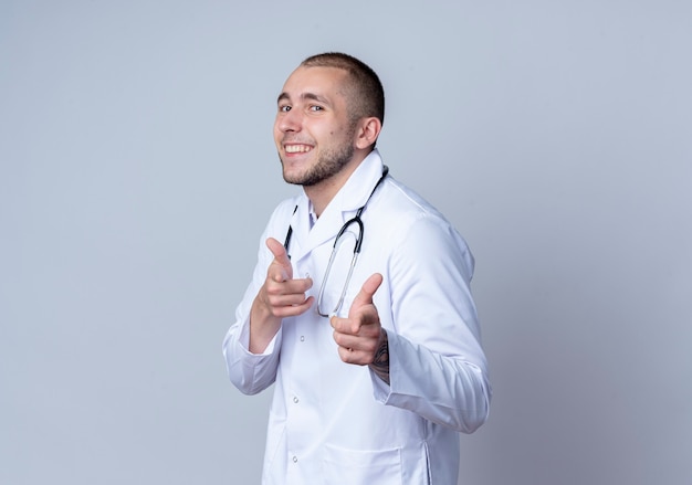 Sonriente joven médico vistiendo bata médica y un estetoscopio alrededor de su cuello haciéndole gesto en la parte delantera aislada en la pared blanca