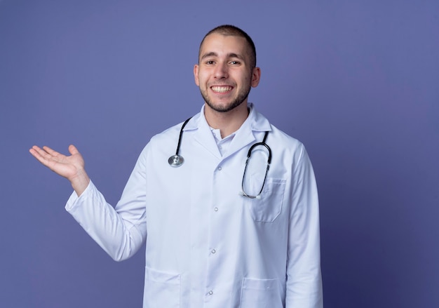 Sonriente joven médico vistiendo bata médica y un estetoscopio alrededor de su cuello mostrando la mano vacía aislada en la pared púrpura
