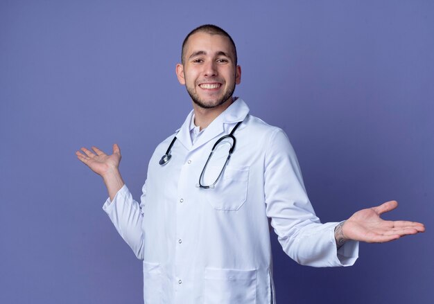 Sonriente joven médico vistiendo bata médica y estetoscopio alrededor de su cuello mostrando las manos vacías aisladas en la pared púrpura