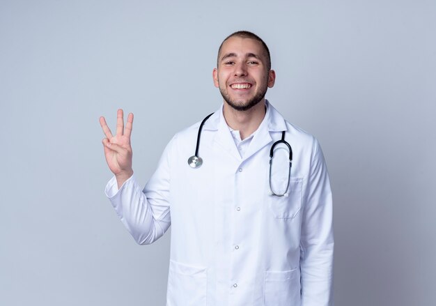 Sonriente joven médico vistiendo bata médica y un estetoscopio alrededor de su cuello mostrando tres con la mano aislada en la pared blanca