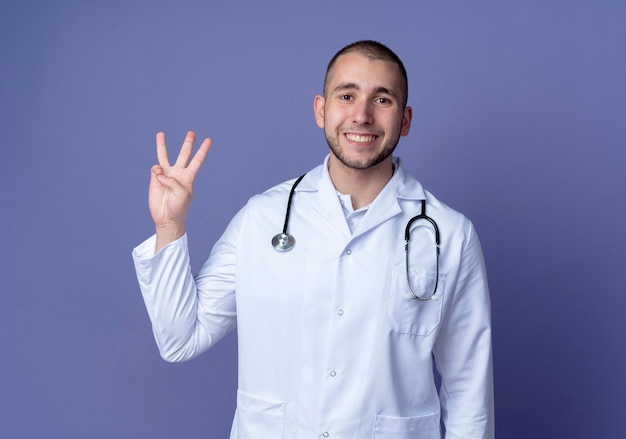 Sonriente joven médico vistiendo bata médica y un estetoscopio alrededor de su cuello mostrando tres con la mano aislada en la pared púrpura