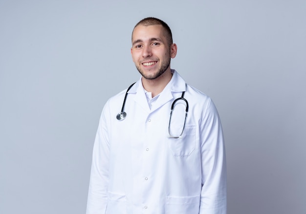 Sonriente joven médico vistiendo bata médica y un estetoscopio alrededor de su cuello de pie y mirando al frente aislado en la pared blanca