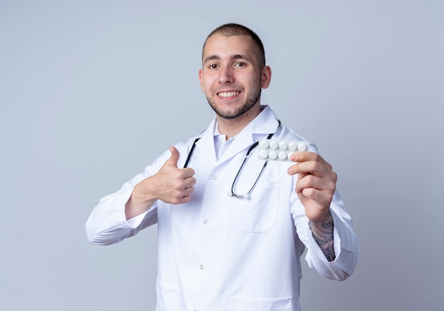 Sonriente joven médico vistiendo bata médica y estetoscopio alrededor de su cuello sosteniendo un paquete de tabletas médicas y mostrando el pulgar hacia arriba aislado en la pared blanca