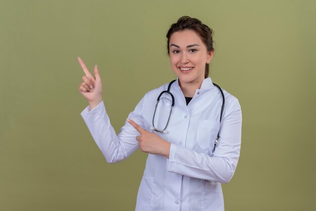 Sonriente joven médico vistiendo bata médica con estetoscopio apunta al lado de la pared verde