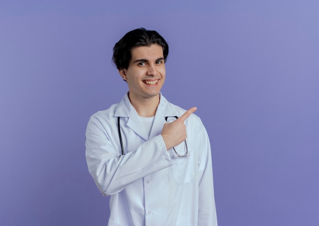 Sonriente joven médico vistiendo bata médica y un estetoscopio apuntando al lado aislado en la pared púrpura con espacio de copia