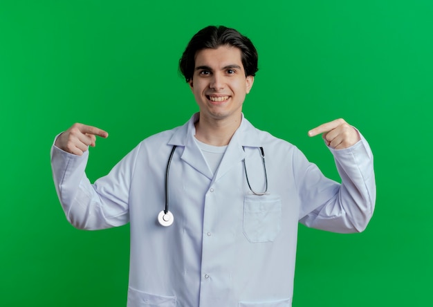 Sonriente joven médico vistiendo bata médica y un estetoscopio apuntando a algo frente a él aislado en la pared verde