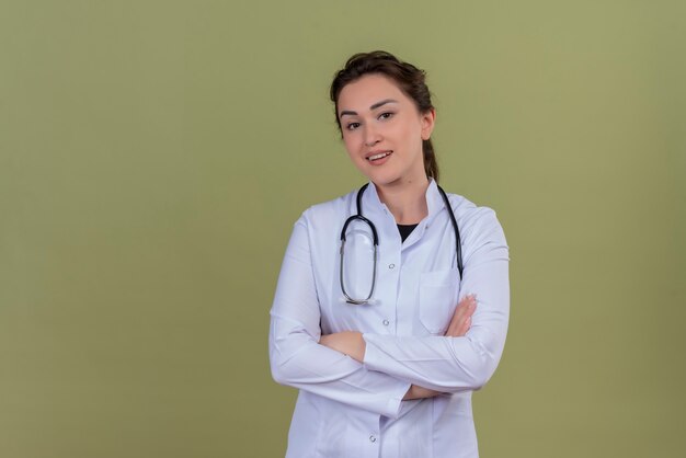 Sonriente joven médico vistiendo bata médica con estetoscopio cruzando las manos sobre la pared verde