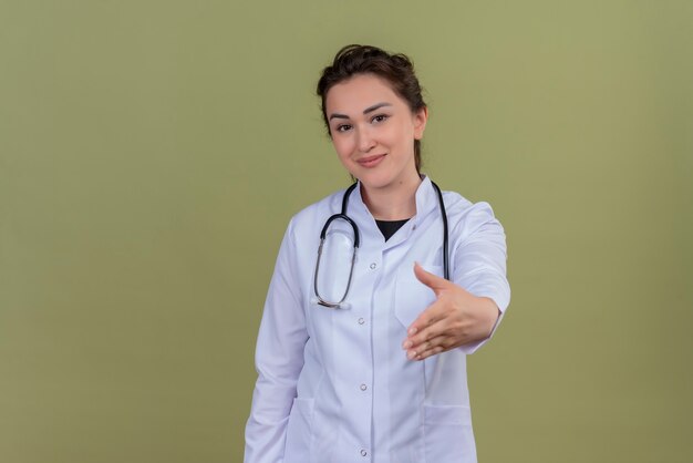 Sonriente joven médico vistiendo bata médica con estetoscopio extendió su mano sobre la pared verde