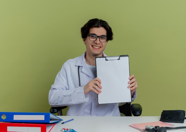 Foto gratuita sonriente joven médico vistiendo bata médica y estetoscopio con gafas sentado en el escritorio con herramientas médicas mirando mostrando portapapeles aislado
