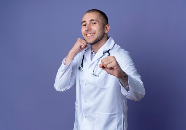 Sonriente joven médico vistiendo bata médica y un estetoscopio haciendo gesto de boxeo en la parte delantera aislada en la pared púrpura